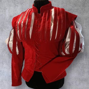 Puede incluir: Un jubón de terciopelo rojo con detalles blancos y mangas abullonadas, exhibido en un maniquí. El corpiño presenta paneles blancos verticales y un frente con cordones. Las mangas son rojas con rayas blancas. Parte de una colección de disfraces.