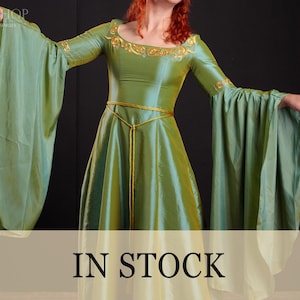Peut inclure: Une robe verte de style médiéval avec de longues manches fluides. La robe présente des broderies dorées autour de l'encolure et une ceinture en corde dorée. Les mots "IN STOCK" sont affichés en bas de l'image.