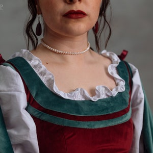 Op de afbeelding: Een close-up van een kostuum in renaissancestijl. Het lijfje is gemaakt van dieprood fluweel, geaccentueerd met teal groene afwerking en witte gerimpelde stof. Een parelketting en donkerrode oorbellen maken de look compleet. Het kostuum is van A l'Entrada Shop.