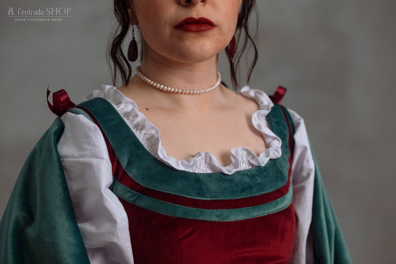 Op de afbeelding: Een close-up van een kostuum in renaissancestijl. Het lijfje is gemaakt van dieprood fluweel, geaccentueerd met teal groene afwerking en witte gerimpelde stof. Een parelketting en donkerrode oorbellen maken de look compleet. Het kostuum is van A l'Entrada Shop.