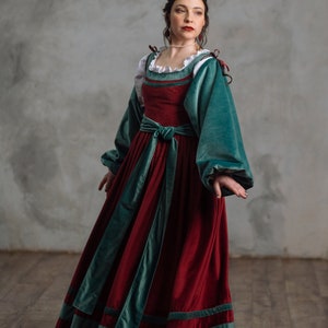 Op de afbeelding: Een bordeauxrode fluwelen jurk met zeegroene accenten. De jurk heeft lange mouwen, een getailleerde taille en een witte gerimpelde halslijn. De jurk komt uit de winkel "A l'eptrada SHOP STAGE COSTUMES & PROPS".