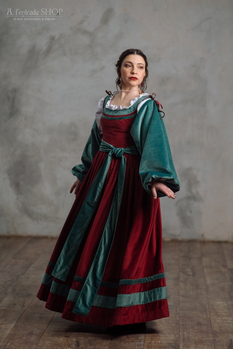 Op de afbeelding: Een bordeauxrode fluwelen jurk met zeegroene accenten. De jurk heeft lange mouwen, een getailleerde taille en een witte gerimpelde halslijn. De jurk komt uit de winkel "A l'eptrada SHOP STAGE COSTUMES & PROPS".