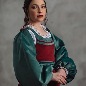 Op de afbeelding: Een teal en bordeaux fluwelen jurk in renaissancestijl met pofmouwen en een witte kraag met ruches. De jurk heeft een getailleerd lijfje en een volle rok. Het model draagt een parelketting.