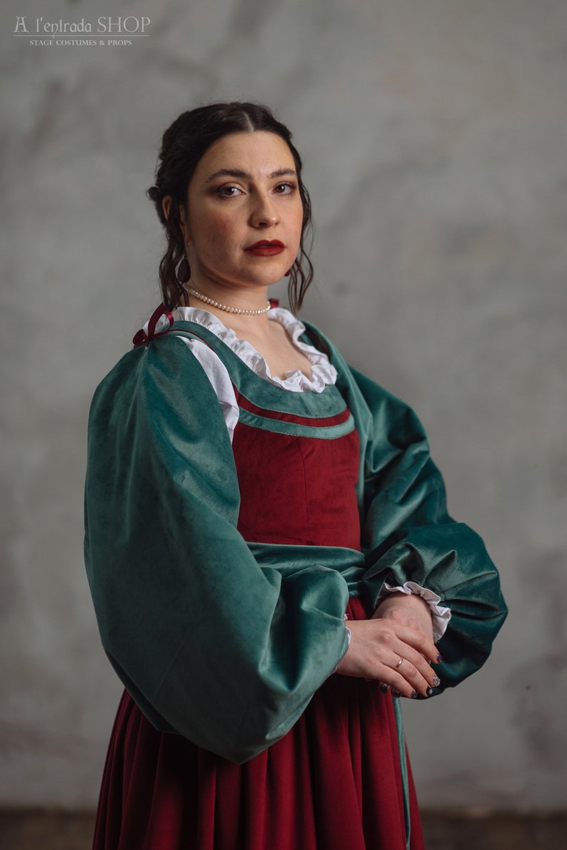 Op de afbeelding: Een teal en bordeaux fluwelen jurk in renaissancestijl met pofmouwen en een witte kraag met ruches. De jurk heeft een getailleerd lijfje en een volle rok. Het model draagt een parelketting.