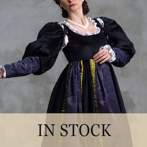 May include: A black and navy blue Renaissance-style dress with puffed sleeves and a full skirt. The dress features gold trim and white lace accents. The text "IN STOCK" is displayed at the bottom of the image.