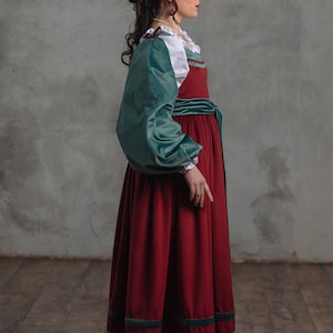 Op de afbeelding: Een lange, rode fluwelen jurk met teal groene accenten en mouwen. De jurk heeft een witte blouse met een gerimpelde kraag en een teal groene fluwelen riem. De tekst "A l'entrada SHOP STAGE COSTUMES & PROPS" is inbegrepen.