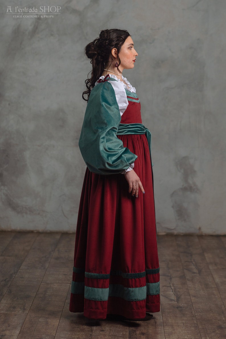 Op de afbeelding: Een lange, rode fluwelen jurk met teal groene accenten en mouwen. De jurk heeft een witte blouse met een gerimpelde kraag en een teal groene fluwelen riem. De tekst "A l'entrada SHOP STAGE COSTUMES & PROPS" is inbegrepen.