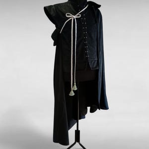 Puede incluir: Un disfraz negro exhibido en un maniquí. El disfraz incluye una chaqueta de cuello alto con una corbata blanca trenzada y borlas, y una capa larga. El texto "A l'entrada SHOP COSTUMES & ACCESSORIES" es visible en la parte superior.