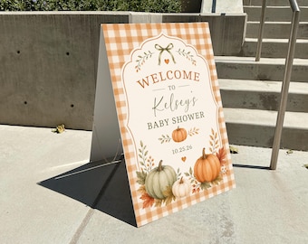 Puede incluir: Un letrero triangular tipo caballete con un patrón de cuadros naranja y blanco. El centro muestra el texto WELCOME TO Kelsey's BABY SHOWER 10.25.26, decorado con ilustraciones de calabazas, hojas de otoño y un pequeño lazo verde arriba.