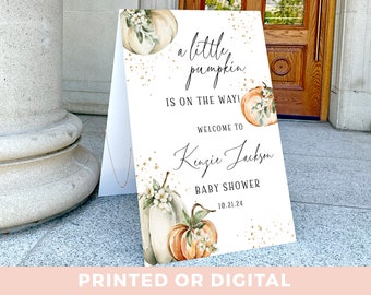 May include: A white sign with gold glitter and a watercolor pumpkin design. The text reads "a little pumpkin is on the way!"  "Welcome to Kenzie Jackson Baby Shower 10.21.24"  "Printed or Digital"