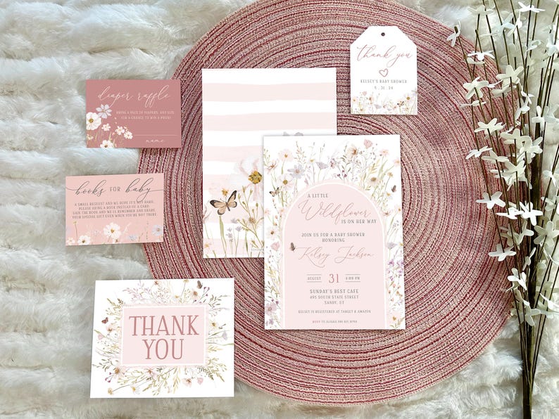 May include: A set of baby shower stationery including an invitation, diaper raffle card, books for baby card, and thank you tags. The cards feature a soft pink and white color palette with watercolor wildflower illustrations, butterflies, and elegant script text.