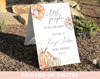 May include: A white sign with gold glitter accents announcing a baby shower for Kenzie Jackson. The sign features a watercolor illustration of pumpkins and flowers with the text "a little pumpkin is on the way!" and "Welcome to Kenzie Jackson Baby Shower 10.21.24".