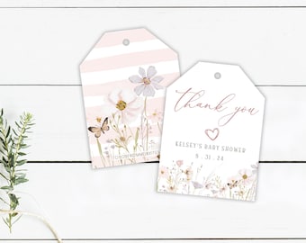 Può includere: Due etichette regalo per baby shower con disegni floreali e farfalle. Un'etichetta ha strisce rosa e un fiore, l'altra dice "Grazie" con un cuore e i dettagli dell'evento. Le etichette sono su una superficie di legno bianco.