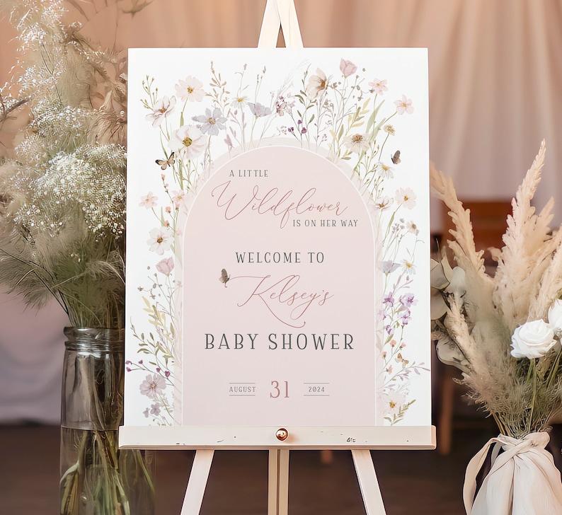 May include: A rectangular baby shower welcome sign featuring a soft pink arch design, watercolor wildflowers, and butterflies. The text reads: A LITTLE Wildflower IS ON HER WAY, WELCOME TO Kelsey's BABY SHOWER, AUGUST 31 2024. The sign is on an easel.