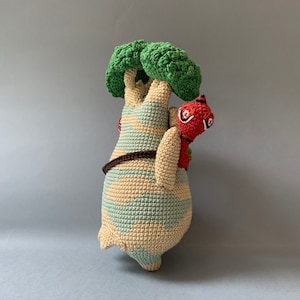 K&ouml;nnte beinhalten: Ein geh&auml;keltes Pl&uuml;schtier, das einen gr&uuml;nen Baum mit braunem Stamm und einem roten und braunen Wesen auf dem R&uuml;cken darstellt. Der Baum hat einen hellblauen und beigefarbenen gestreiften K&ouml;rper.