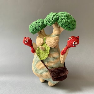 K&ouml;nnte beinhalten: Ein geh&auml;keltes Pl&uuml;schtier, das einem Baum &auml;hnelt, mit einer braunen Tasche und zwei roten, vogelartigen Figuren, die daran befestigt sind. Der Baum ist aus beigefarbenem, gr&uuml;nem und braunem Garn gefertigt.