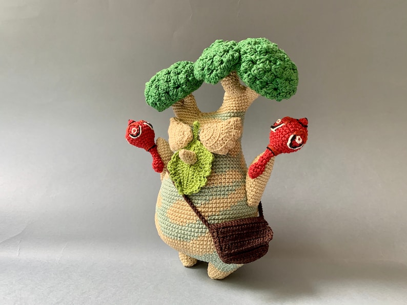 K&ouml;nnte beinhalten: Ein geh&auml;keltes Pl&uuml;schtier, das einem Baum &auml;hnelt, mit einer braunen Tasche und zwei roten, vogelartigen Figuren, die daran befestigt sind. Der Baum ist aus beigefarbenem, gr&uuml;nem und braunem Garn gefertigt.