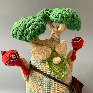 K&ouml;nnte beinhalten: Ein geh&auml;keltes Pl&uuml;schtier in Form eines Baumes mit gr&uuml;nen Bl&auml;ttern und einem braunen Stamm. Der Baum hat zwei rote geh&auml;kelte Arme mit einem roten und wei&szlig;en geh&auml;kelten Kreis an jedem Arm.