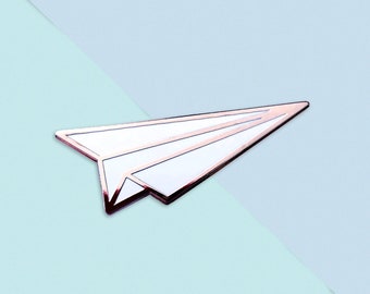 Könnte beinhalten: Eine weiße Papierflieger-Anstecknadel mit roségoldener Umrandung. Die Anstecknadel befindet sich auf einem hellblauen und weißen Hintergrund.