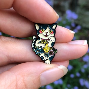 Può includere: Uno spillo in smalto bianco e nero di un gatto che tiene una moneta d'oro con fiori intorno.