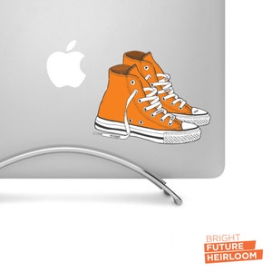May include: Orange high-top sneakers with white laces and a white sole. The sneakers are on a silver laptop stand with a white Apple logo on the laptop.