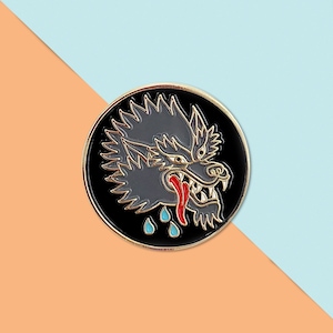 May include: A black and gold enamel pin with a detailed image of a wolf's head with a red tongue and blue teardrop shapes.