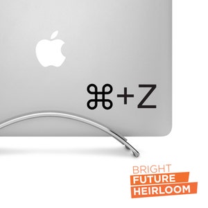 May include: A silver laptop computer with a black Apple logo and a black sticker that says "⌘ + Z" on the top right corner. The laptop is on a silver stand. The text "BRIGHT FUTURE HEIRLOOM" is printed in orange on a white background.