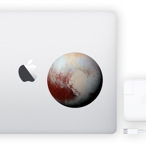 May include: A white laptop with a silver apple logo and a sticker of Pluto, a dwarf planet with a reddish-brown surface and a light blue and white icy cap.