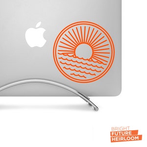 May include: Orange line art sticker of a sun setting over the ocean waves in a circle. The sticker is on a silver laptop.