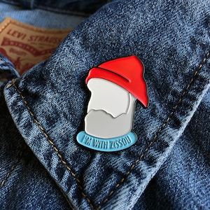 Puede incluir: Un pin de esmalte azul y blanco con una ilustración de dibujos animados de una persona con un sombrero rojo y el texto "I'm with Zissou".