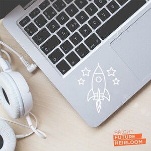 May include: A white laptop computer with a keyboard and a trackpad. The trackpad has a white sticker with a rocket ship and stars on it. The text "BRIGHT FUTURE HEIRLOOM" is printed on the bottom right corner of the image.