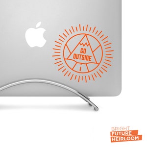 May include: Orange sticker with a mountain and sun design on a silver laptop. The sticker says "GO OUTSIDE".