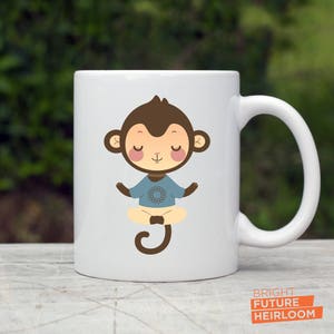 May include: White ceramic mug with a cartoon illustration of a brown monkey meditating in a blue shirt with a mandala design. The monkey has closed eyes and a peaceful expression.