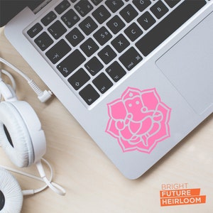 May include: A pink sticker of the Hindu deity Ganesha on a silver laptop. The sticker is a silhouette of Ganesha sitting in a lotus flower. The laptop has a black keyboard and a trackpad.