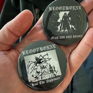 May include: Two black Bloodborne button pins. One pin features a silhouetted figure with the text "FEAR THE OLD BLOOD". The other pin has a creature graphic with the text "Hail The Nightmare".