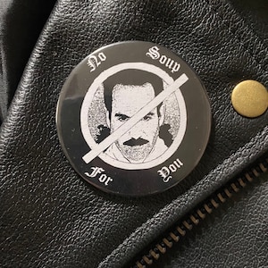 May include: A black and white button with the text "No Soup For You" encircling a portrait of a man with a mustache, crossed out with a white line. The button is on a black leather jacket.