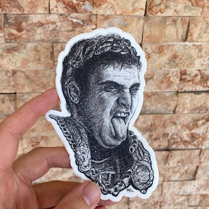 May include: A black and white sticker featuring a detailed illustration of a man with his tongue sticking out. The man is wearing a laurel wreath and ornate clothing. The sticker has a white border and is held by a hand.
