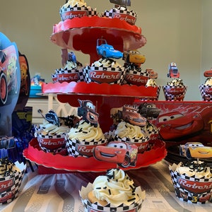 May include: A three-tiered red cupcake stand with white and black checkered cupcake liners. The cupcakes are topped with white frosting and black sprinkles. The cupcakes are decorated with Cars characters, including Lightning McQueen, Mater, and  'Robert 3' on the cupcake liners.