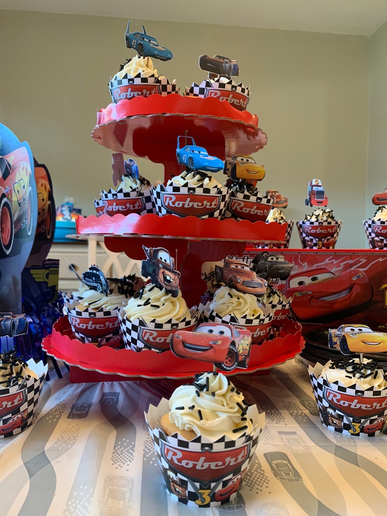 May include: A three-tiered red cupcake stand with white and black checkered cupcake liners. The cupcakes are topped with white frosting and black sprinkles. The cupcakes are decorated with Cars characters, including Lightning McQueen, Mater, and  'Robert 3' on the cupcake liners.