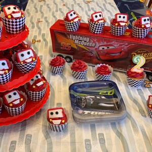 May include: A Cars themed birthday party with red and white checkered cupcakes decorated with Lightning McQueen and Mater characters. There is a red and white tiered cupcake stand, a red toy truck, and a red and black Cars themed plate with a black toy car.