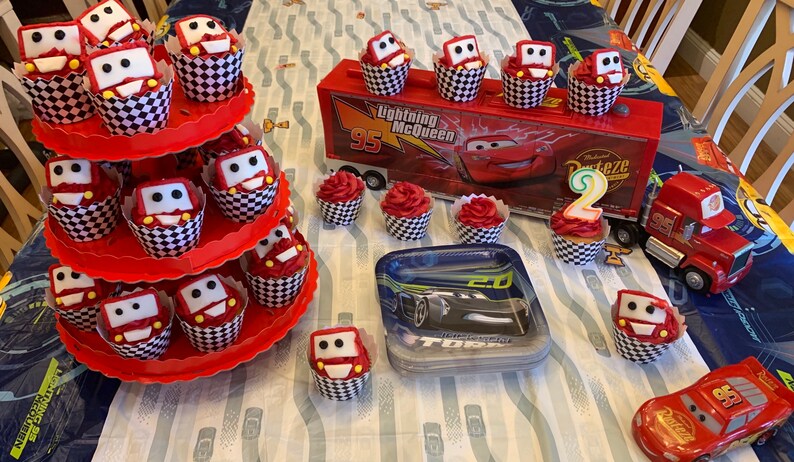 May include: A Cars themed birthday party with red and white checkered cupcakes decorated with Lightning McQueen and Mater characters. There is a red and white tiered cupcake stand, a red toy truck, and a red and black Cars themed plate with a black toy car.
