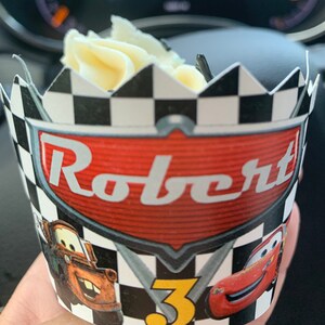May include: A black and white checkered cupcake wrapper with a red, white, and black Cars themed design. The wrapper features the text "Robert" and the number "3".