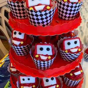 May include: A three-tiered red cupcake stand with black and white checkered cupcake liners. Each cupcake is decorated with red and white frosting and a white fondant car with black eyes and yellow wheels.