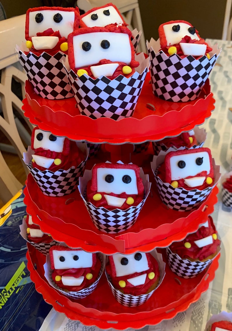 May include: A three-tiered red cupcake stand with black and white checkered cupcake liners. Each cupcake is decorated with red and white frosting and a white fondant car with black eyes and yellow wheels.