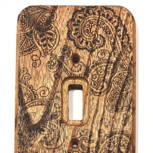 May include: A wooden light switch cover with a paisley design burned into the surface. The wood is a dark brown color with a natural grain pattern.