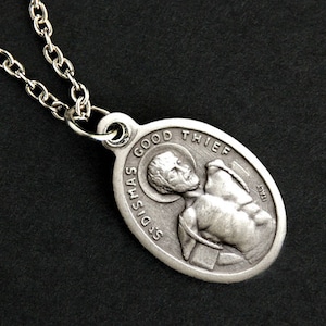 Może przedstawiać: Srebrny łańcuszek z srebrnym, owalnym wisiorkiem przedstawiającym płaskorzeźbę mężczyzny z napisem "St. Dismas Good Thief" wokół krawędzi.