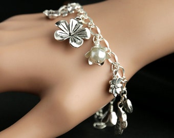 May include: A silver charm bracelet featuring flower and leaf charms. The bracelet has a chain link design and includes a pearl charm. The bracelet is worn on a wrist.