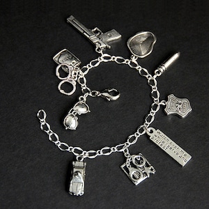 May include: Silver charm bracelet with various police-themed charms, including a gun, handcuffs, sunglasses, a police badge, a bullet, a fingerprint, a police car, and a charm with the text "Watch over me while I serve and protect".