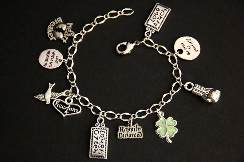 May include: A silver charm bracelet with multiple charms, including a heart with the word "freedom" on it, a dove, a four-leaf clover, a boxing glove, and several rectangular charms with phrases like "Live with no regrets", "Laugh often", "Happily Divorced", "Love much", and "Be true to yourself".