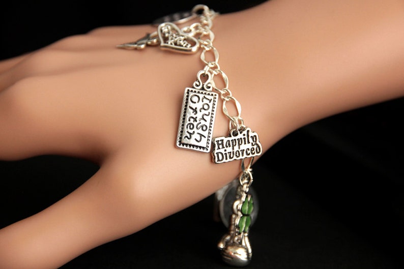 May include: Silver charm bracelet with multiple charms, including a heart charm with the word "Love" on it, a charm with the words "Laugh Often", a charm with the words "Happily Divorced", and a green shamrock charm.